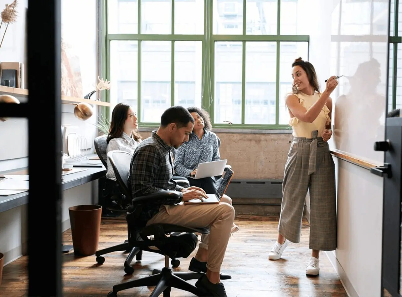 A group of colleagues in a bright office space with one person writing on a whiteboard and others discussing work on a laptop.