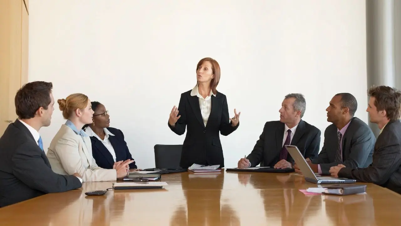 A woman standing and presenting at a business meeting while a group of colleagues listens.