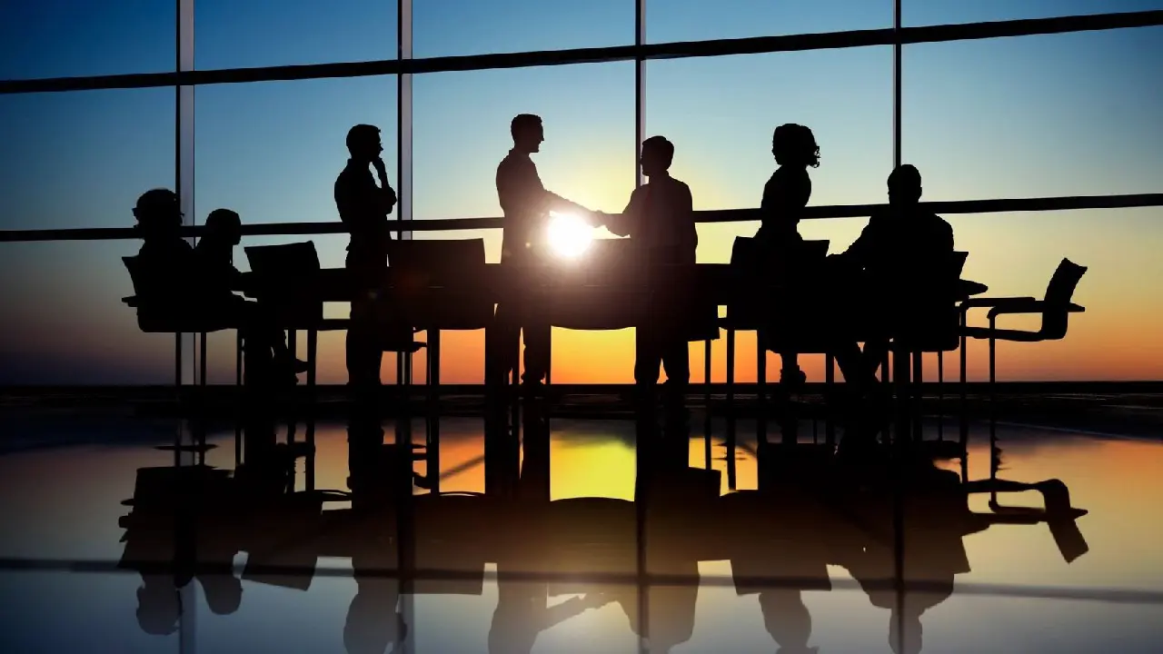 Business professionals shaking hands during a meeting at sunset, silhouetted against an office window.