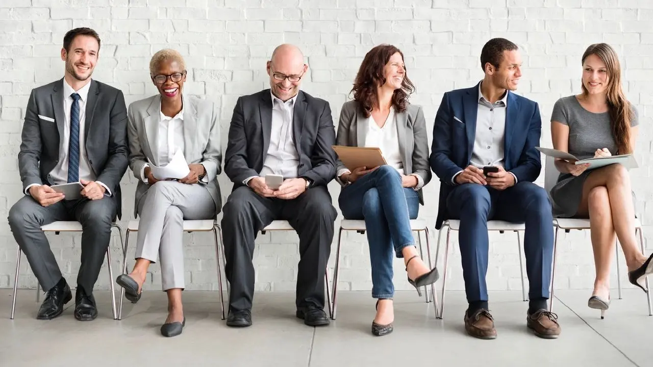 Six professionals smiling and sitting in a row on chairs against a white brick wall, some holding tablets and papers.