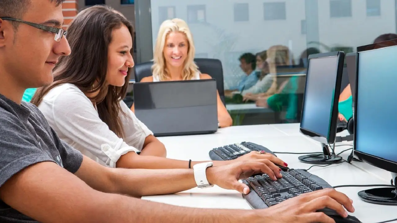 Office employees working on computers while a colleague looks on from the background.