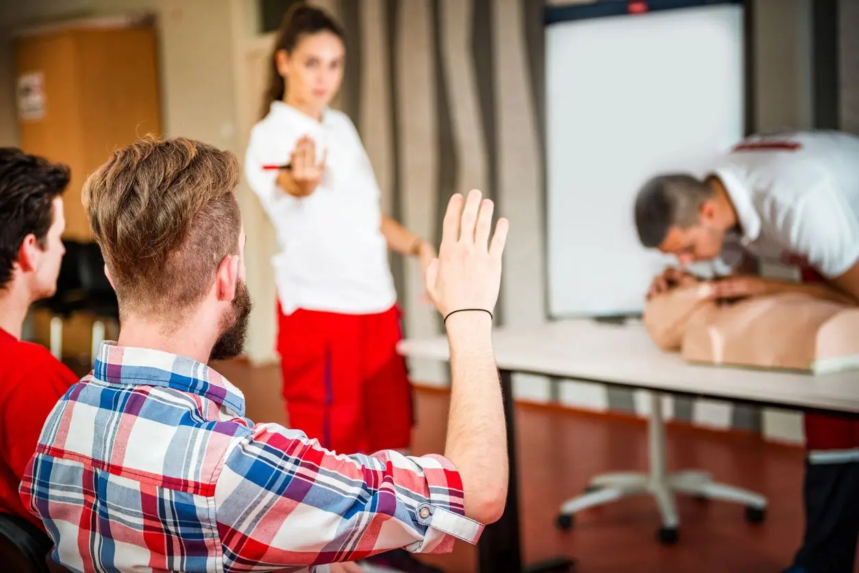 A student raises a hand to ask a question during a cpr training class while an instructor and another student practice on a mannequin.