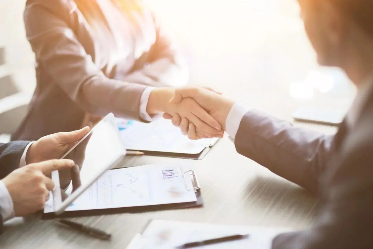 Two professionals engaging in a handshake over a business meeting desk with documents and a digital tablet.